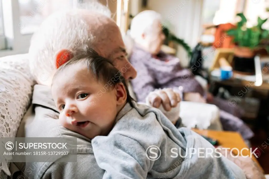 Great-grandfather holding baby at home with his wife in background