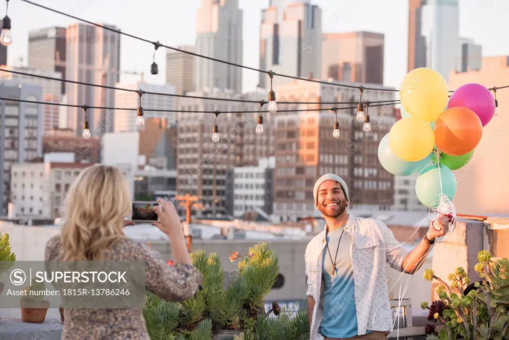 Young couple with balloons standing on rooftop terrace, using smart phone