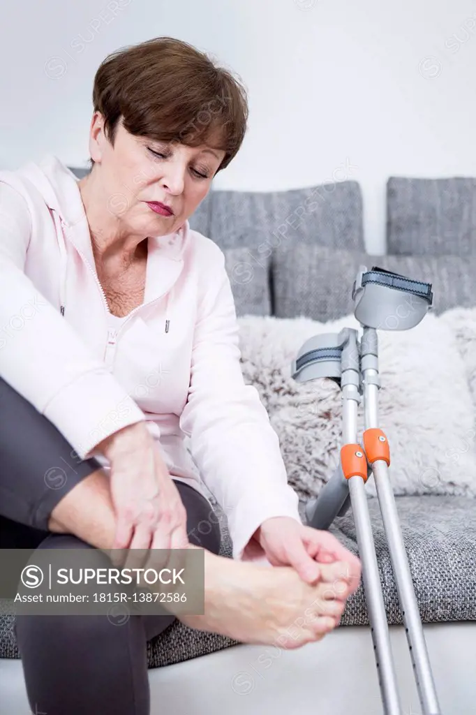 Senior woman with crutches sitting on couch, checking her ankle