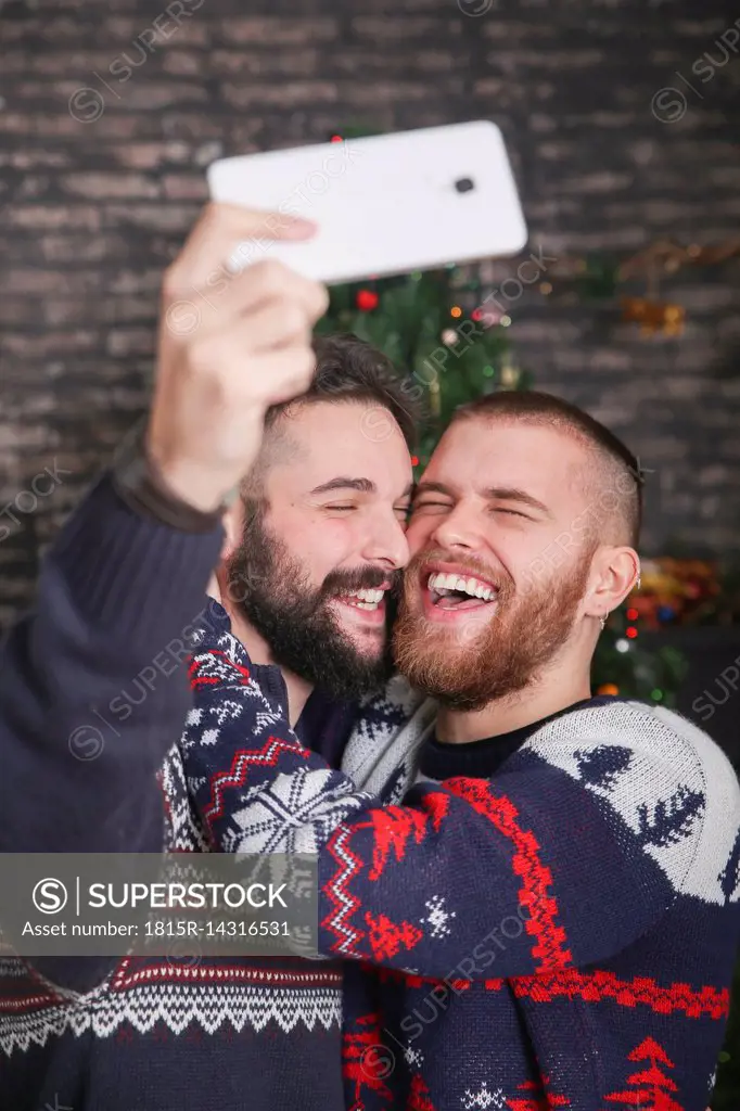 Portrait of laughing gay couple taking selfie with smartphone at Christmas time at home