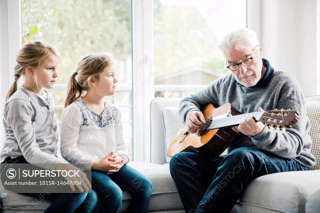 Two girls sitting on sofa listening to grandfather playing guitar