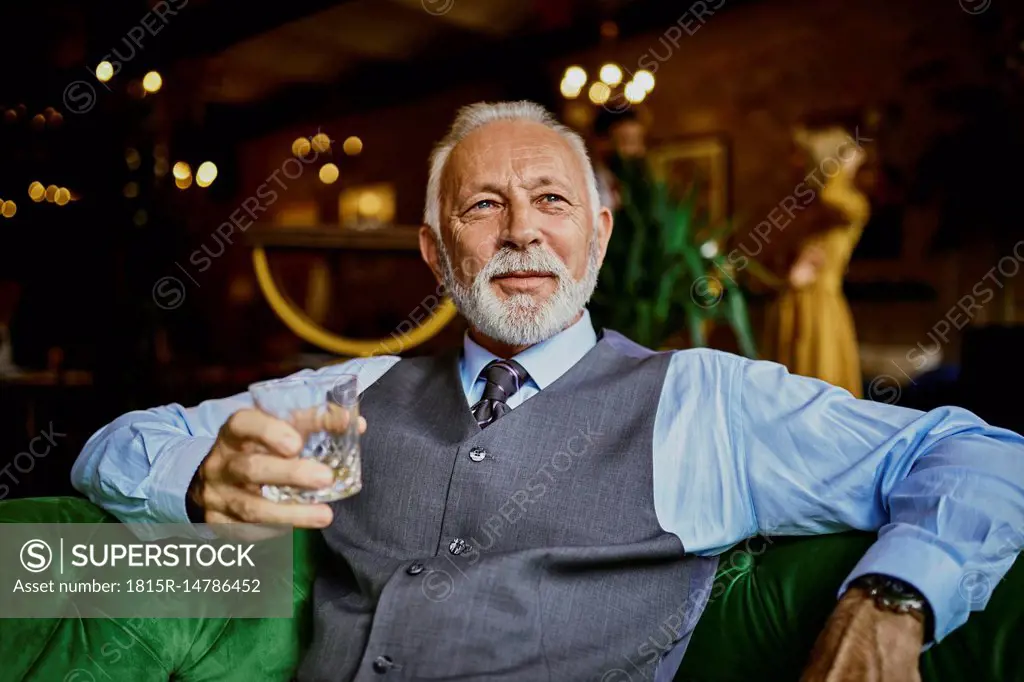 Portrait of elegant senior man sitting on couch in a bar holding tumbler