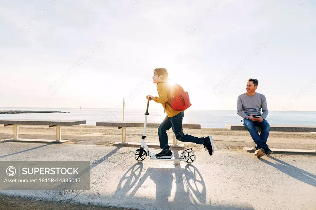 Father watching son riding scooter on beach promenade at sunset