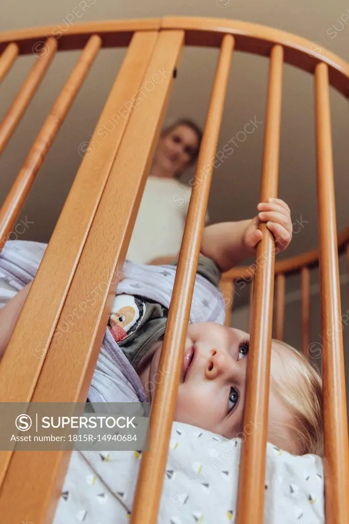 Baby lying in crib with open eyes, mother watching in background