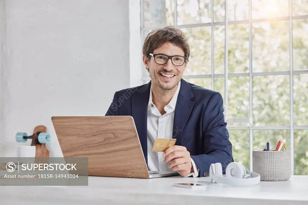 Portrait of smiling businessman with laptop on desk holding credit card