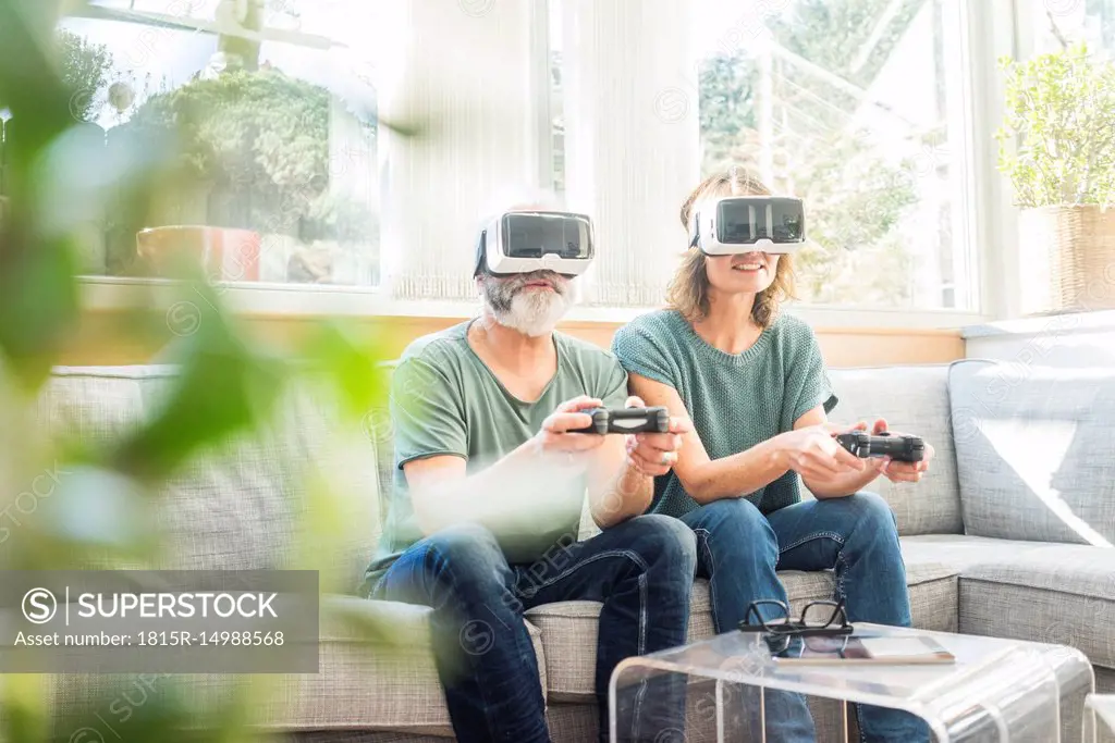 Mature couple sitting on couch at home wearing VR glasses playing video game