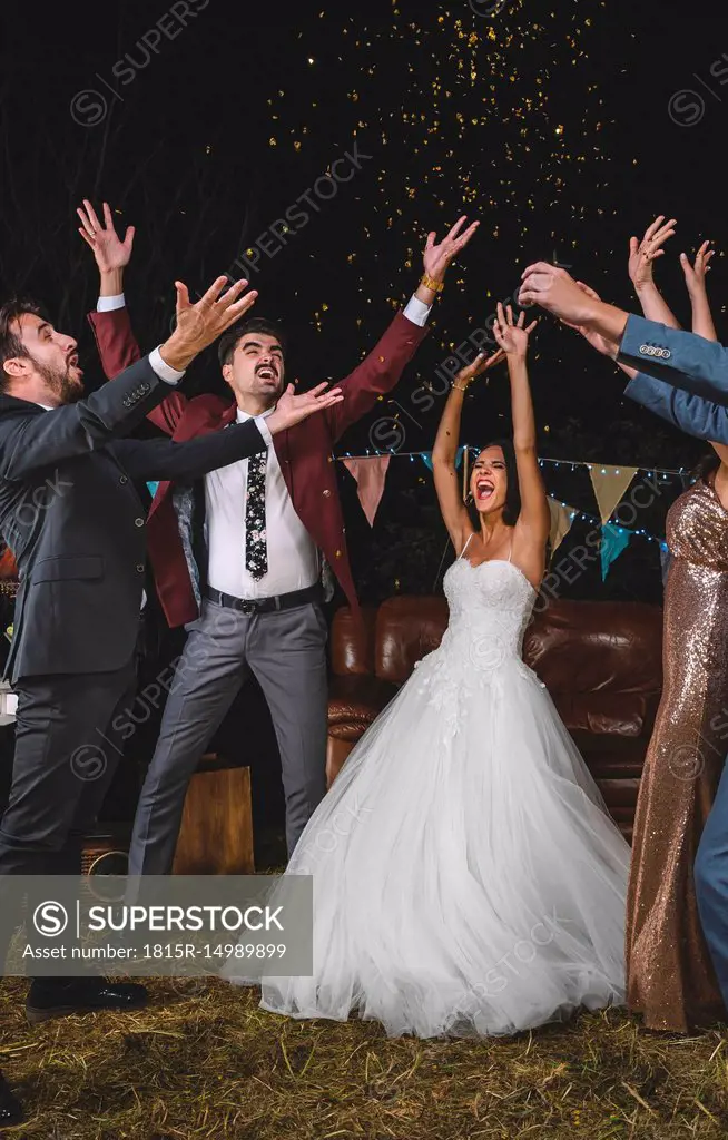 Cheerful wedding couple and friends raising their arms while confetti falling over their heads on a night party outdoors