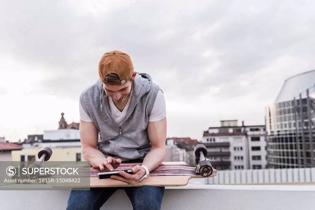 Smiling man sitting in the city with skateboard using cell phone