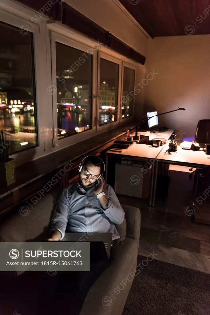 Businessman in office sitting on the couch at night using laptop and headphones