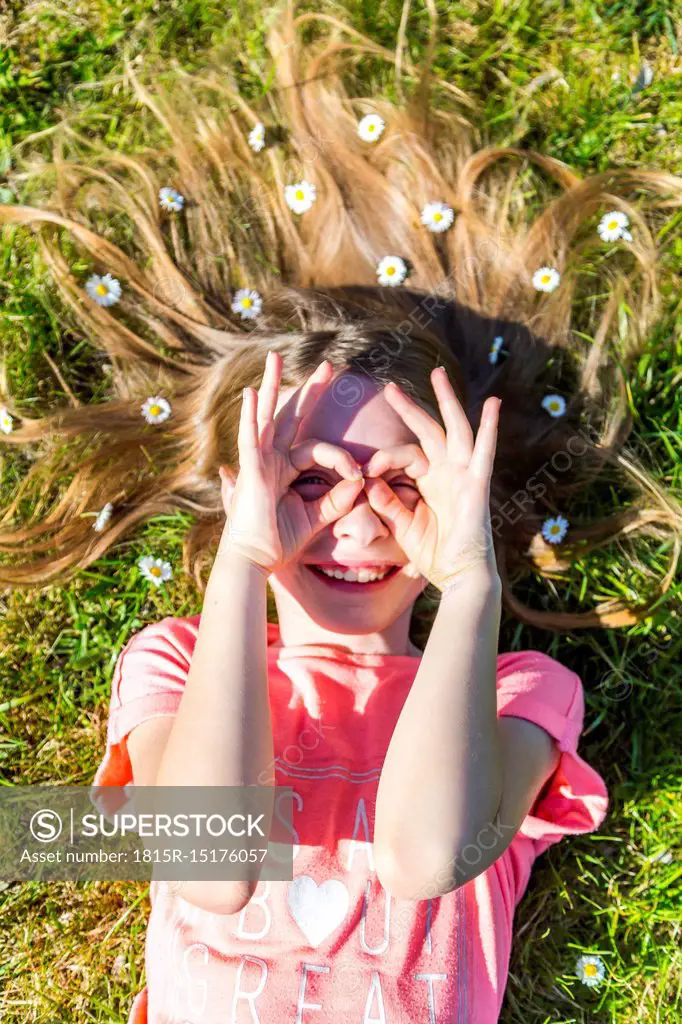 Smiling girl lying on grass in spring with daisies on hair forming spectacles with fingers