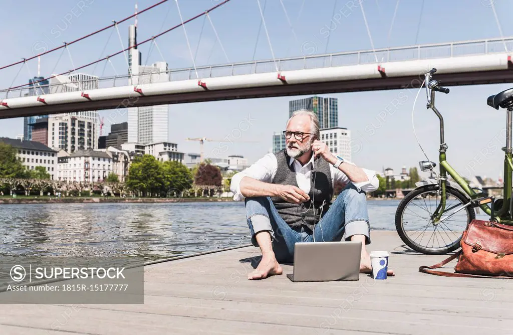 Mature man with laptop, earbuds and bicycle sitting at the riverside in the city