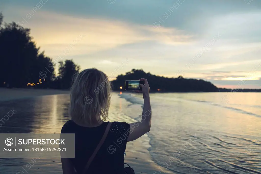 Thailand, Khao Lak, back view of woman taking photo with cell phone on the beach at sunset