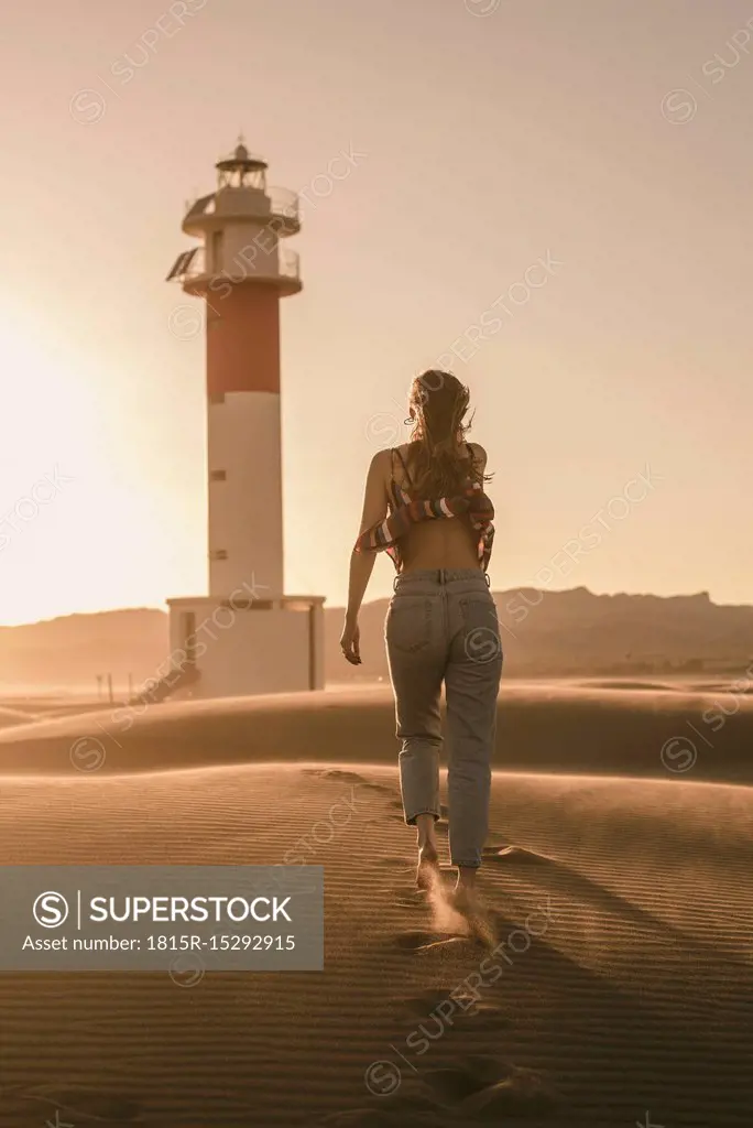 Spain, back view of young woman walking to lighthouse at sunset