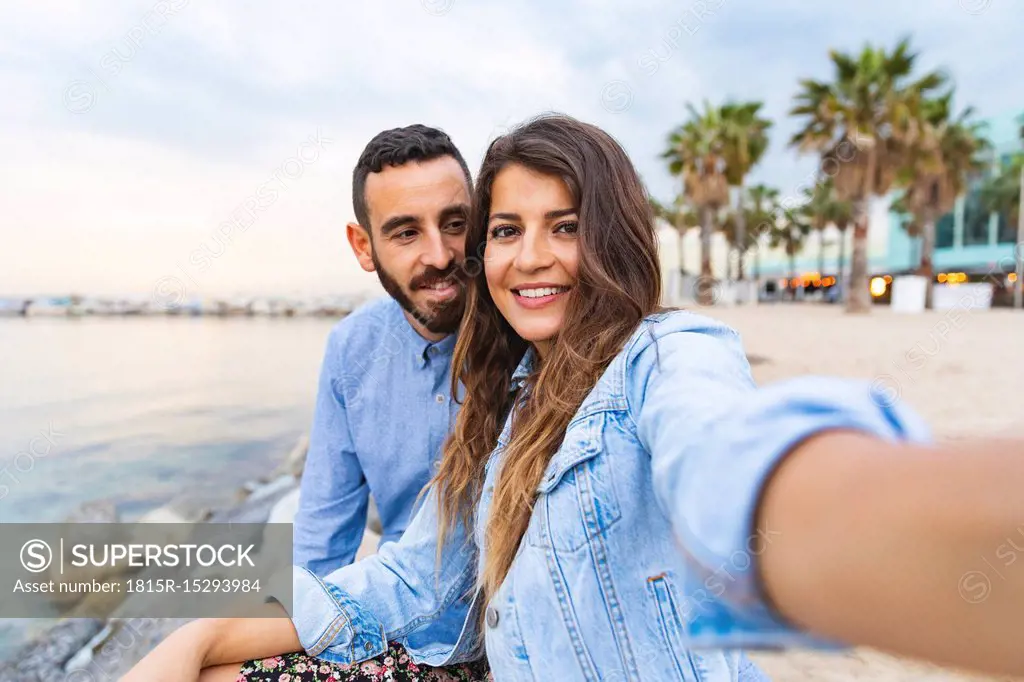 Spain, Barcelona, smiling couple taking a selfie at the seaside