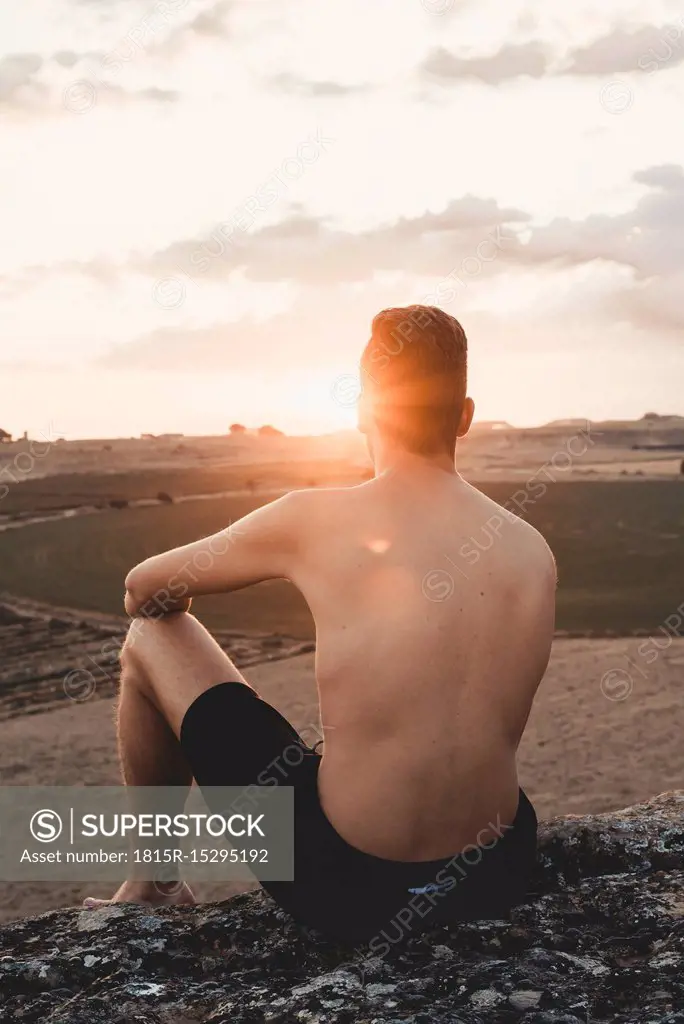 Shirtless young man sitting on rock, watching sunset