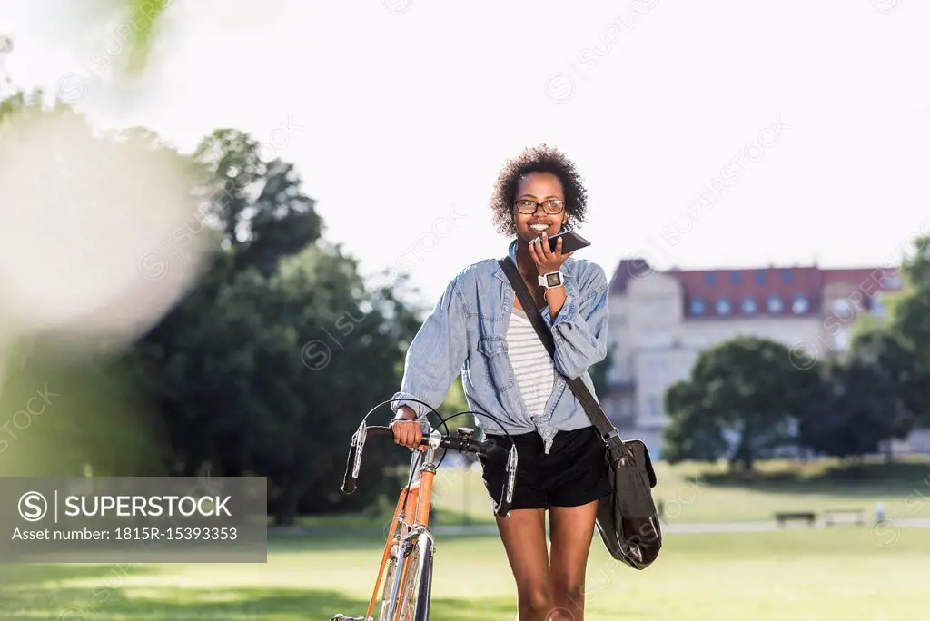 Smiling young woman with cell phone and bicycle in park