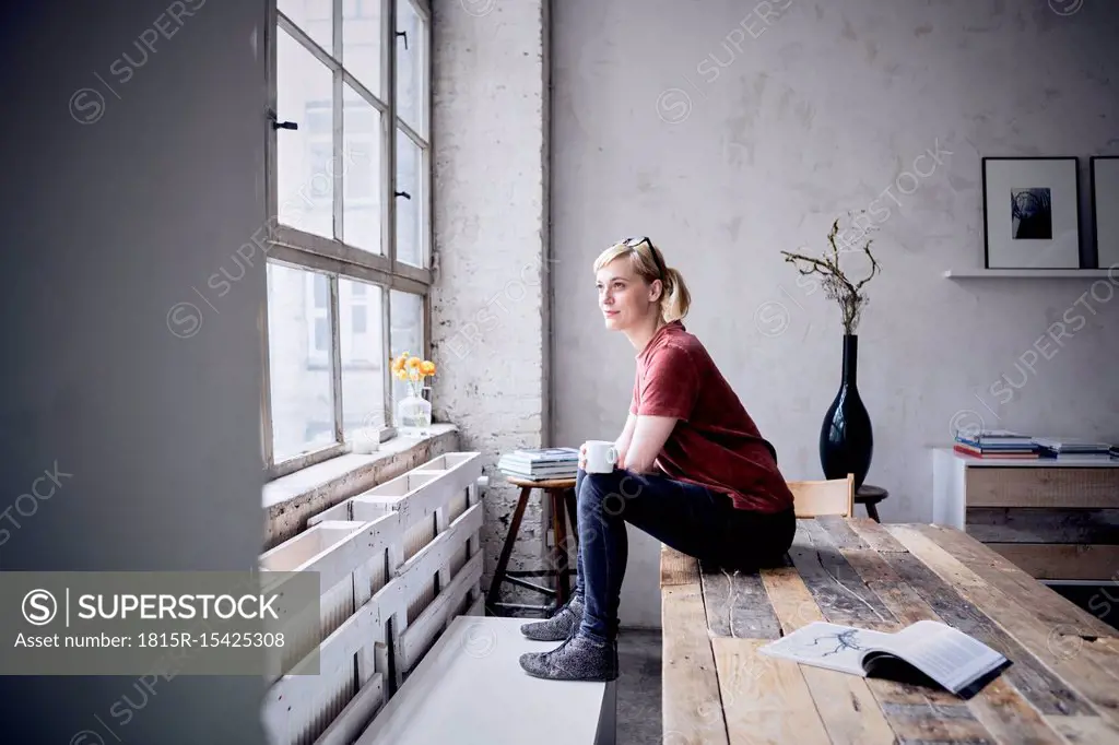 Smiling woman with coffee mug sitting on desk in loft looking through window