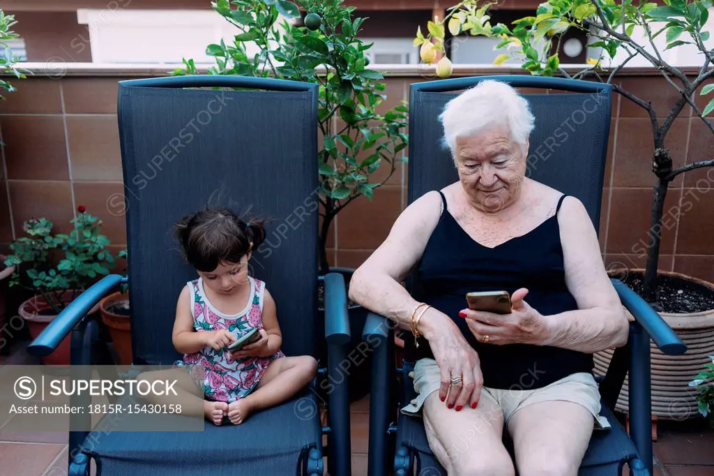 Grandmother and baby girl sitting side by side on the terrace using mobile phones