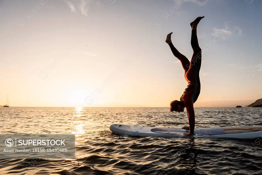 Young man doing handstand on paddleboard at sunset