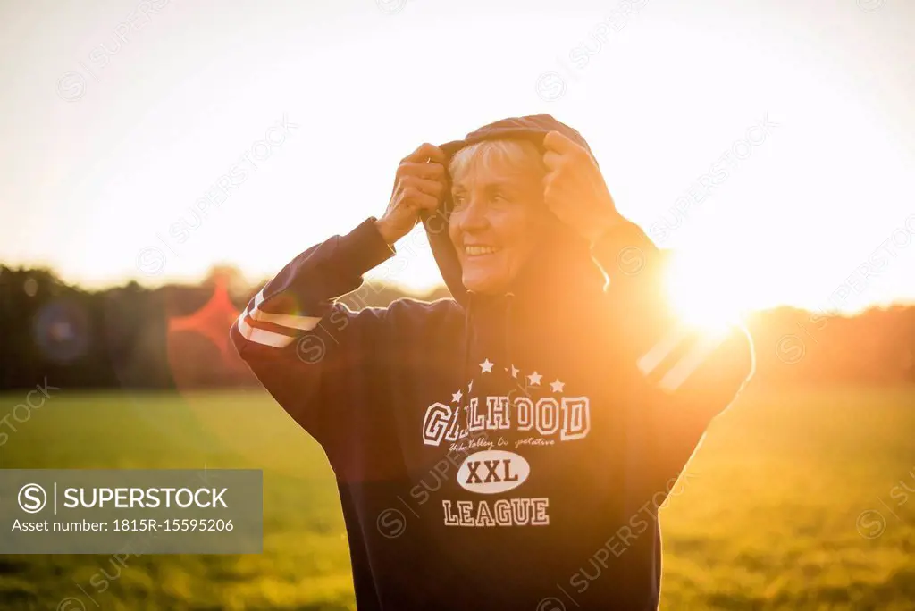 Smiling senior woman wearing a hoodie standing on rural meadow at sunset