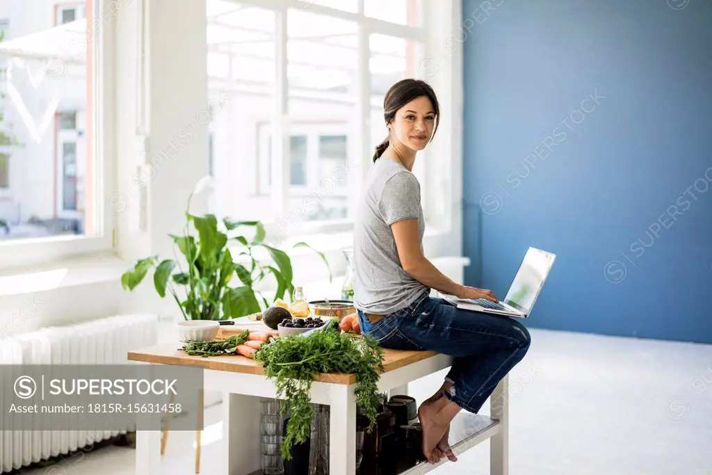 Woman sitting on kitchen table, searching for healthy recipes, using laptop