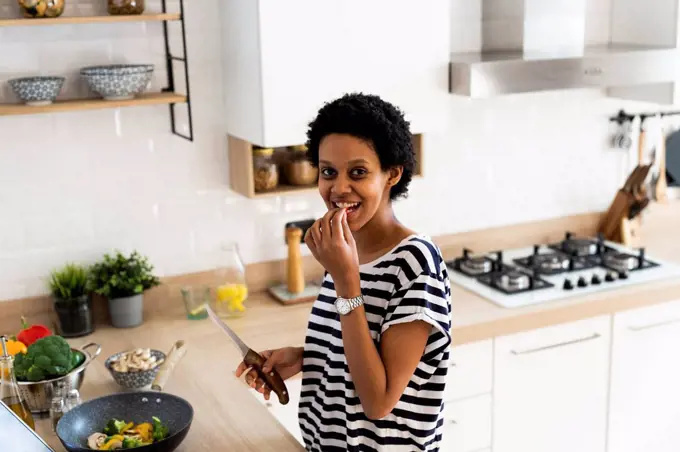 Portrait of smiling young woman cooking in kitchen at home tasting vegetables