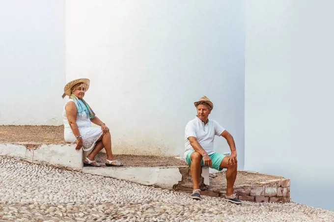 Senior tourist couple sitting on steps in a village, El Roc de Sant Gaieta, Spain