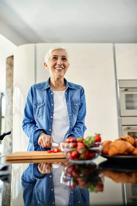 Senior woman standing in kitchen, chopping strawberries