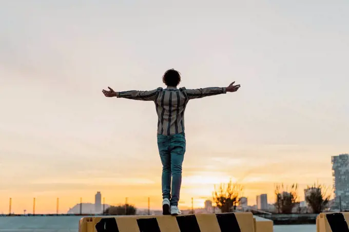 Back view of man with arms outstretched standing on barrier enjoying sunset, Barcelona, Spain