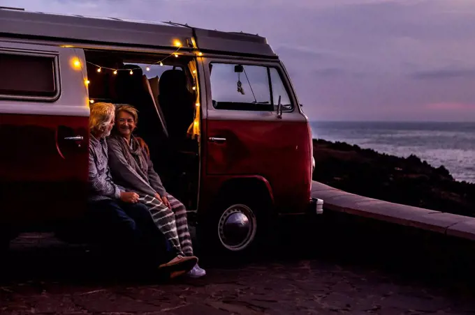 Senior couple traveling in a vintage van, watching sunset at the sea