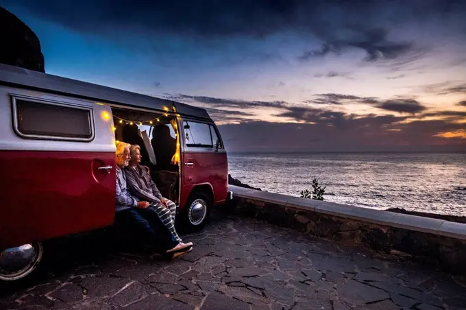 Senior couple traveling in a vintage van, watching sunset at the sea