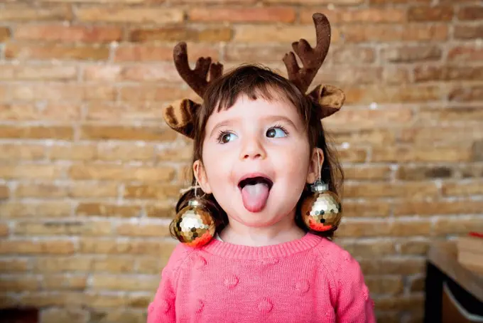 Portrait of toddler girl sticking out tongue wearing reindeer antlers headband and Christmas baubles