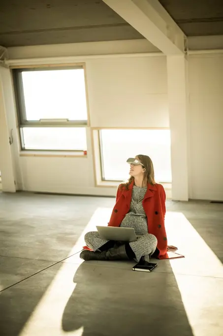 Pregnant busnesswoman sitting on floor of new office rooms, using VR goggles and laptop