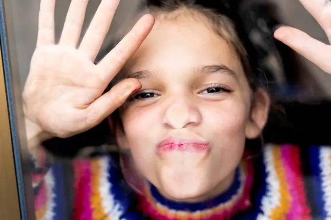 Portrait of happy playful girl in striped pullover behind windowpane