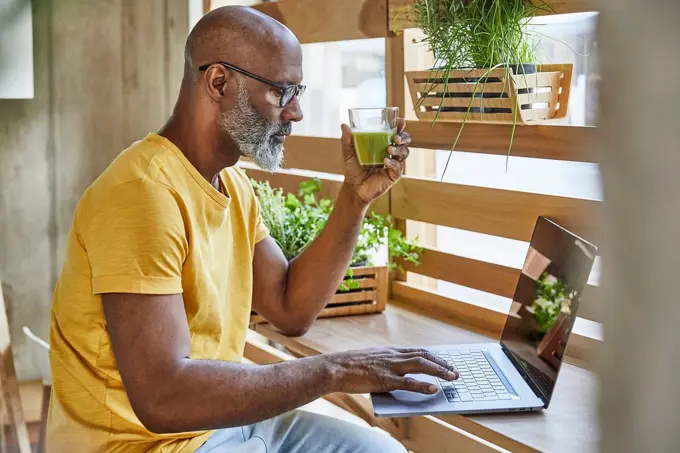 Mature businessman with smoothie using laptop at the window in office