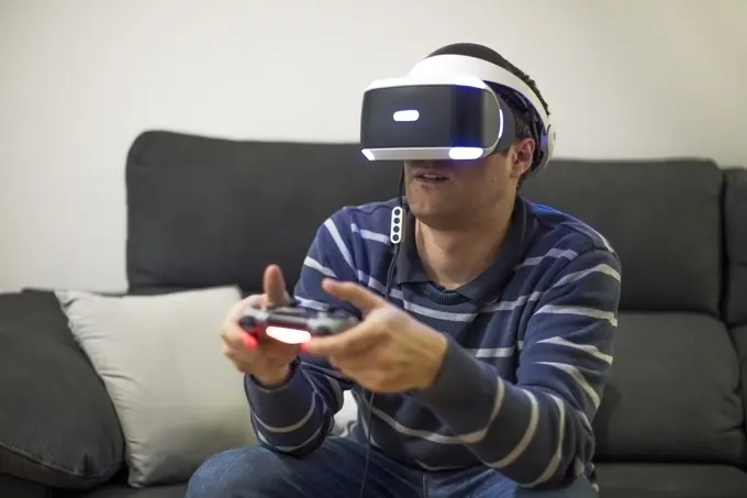 Young man wearing VR glasses sitting on couch at home playing video game