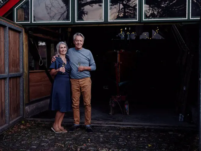 Senior couple standing in front of boathouse with glass of champagne