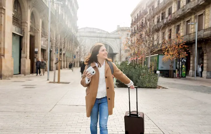 Smiling young woman with suitcase and camera in the city on the go, Barcelona, Spain