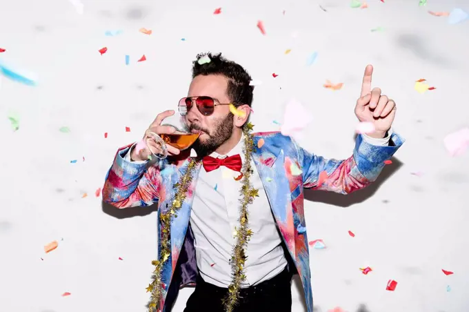 Cool and stylish man wearing a colorful suit and sunglasses celebrating a party with confetti and drinking