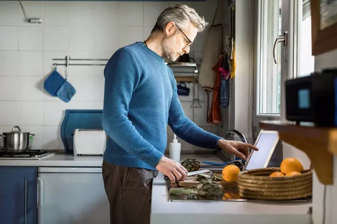 Mature man preparing artichokes in his kitchen using digital tablet