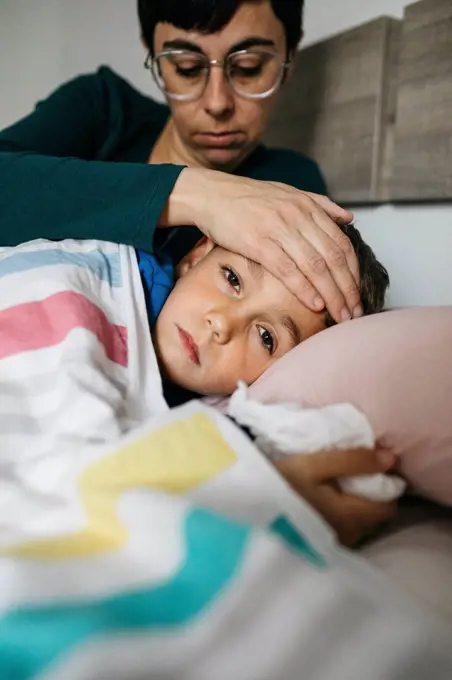 Portrait of sick boy lying in bed while his mother touching his forehead