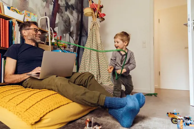 Son tying his father working on laptop in children's room