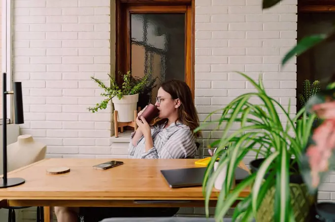 Thoughtful woman drinking coffee while sitting at desk in home office