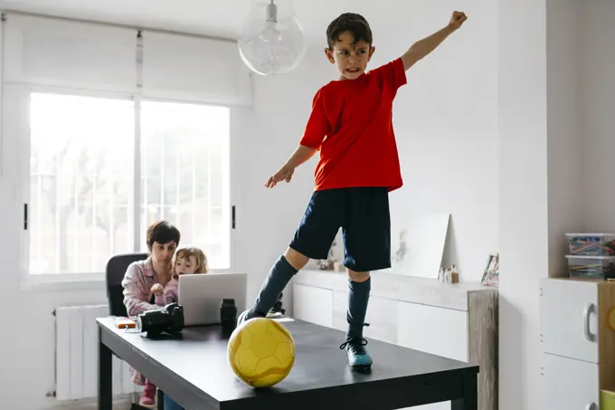 Mom working at home with her children, son standing on soccer ball on table raising arm