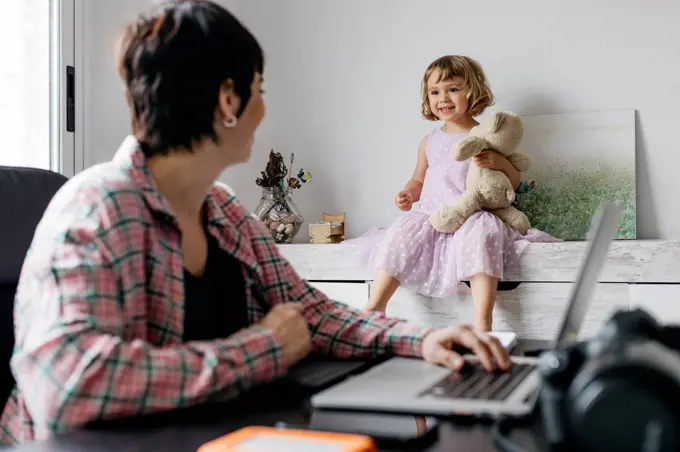 Mother working at home, daughter sitting with teddy bear on sideboard