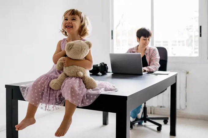 Mother working at home, daughter sitting with teddy bear on table