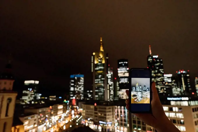 Germany, Hesse, Frankfurt, Hands of woman taking smart phone photos of city downtown at night