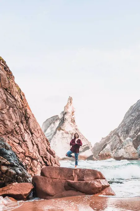 Young woman standing on rock, tree position, Praia da Ursa, Lisboa, Portugal