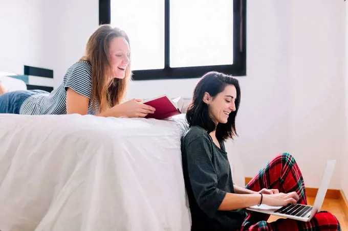 Smiling woman sitting on the floor using laptop while her friend watching from bed