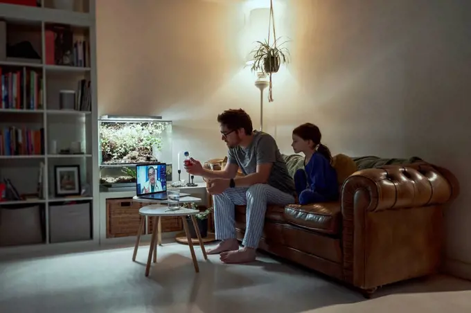 Sick girl sitting by father discussing telemedicine with doctor over video call through laptop in living room at home